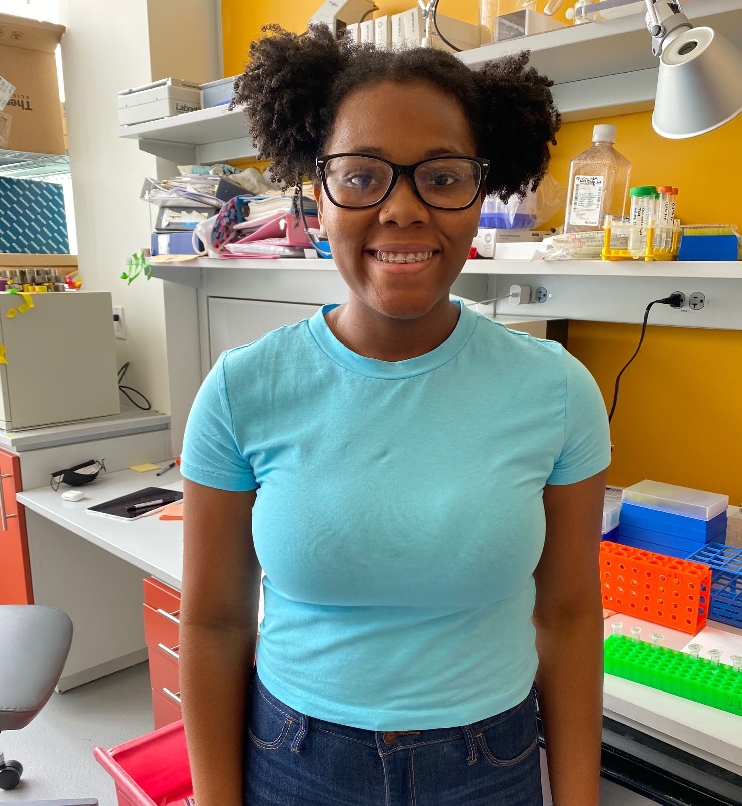 Joanne is wearing a teal tee and standing in front of her bench, with colorful eppendorf racks filled with tubes.