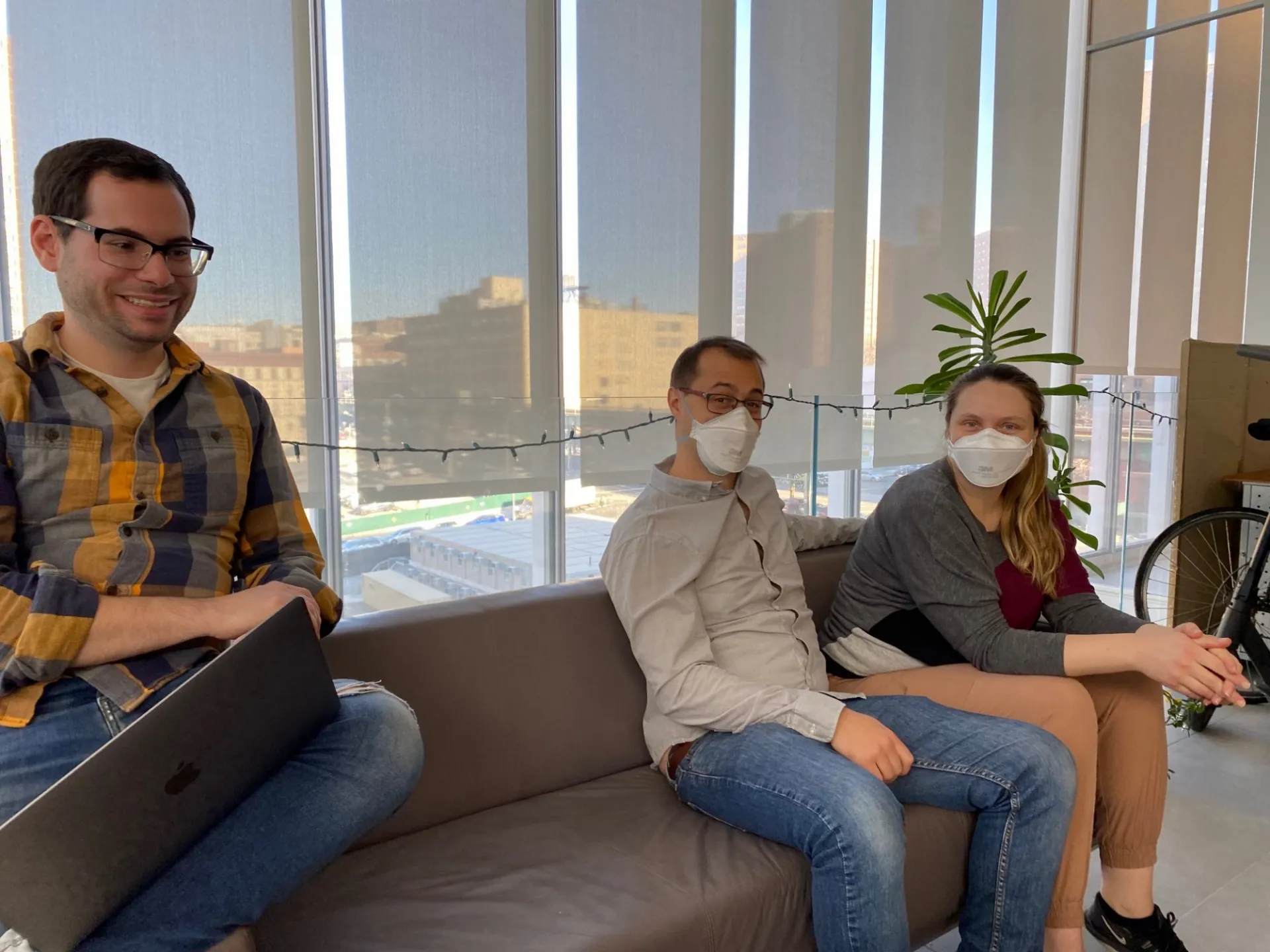 Hani Shayya is wearing yellow and blue plaid and is at his laptop on the couch. Further on the couch, Marco is seated in a beige button up and Lisa is wearing a sweater. 
