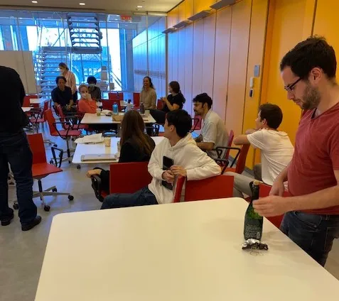 Hani is wearing a red tee and opening a bottle of champagne. A dozen lab members are seated at small tables and looking around the room.