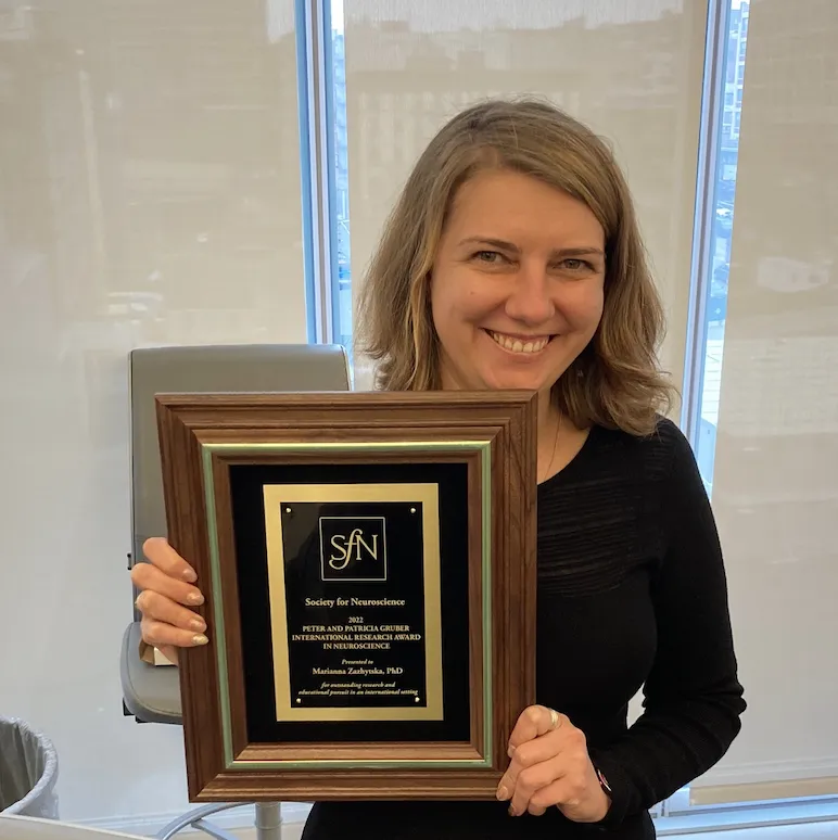 Marianna is smiling and holding up her plaque award that has the "SfN" logo and her name.