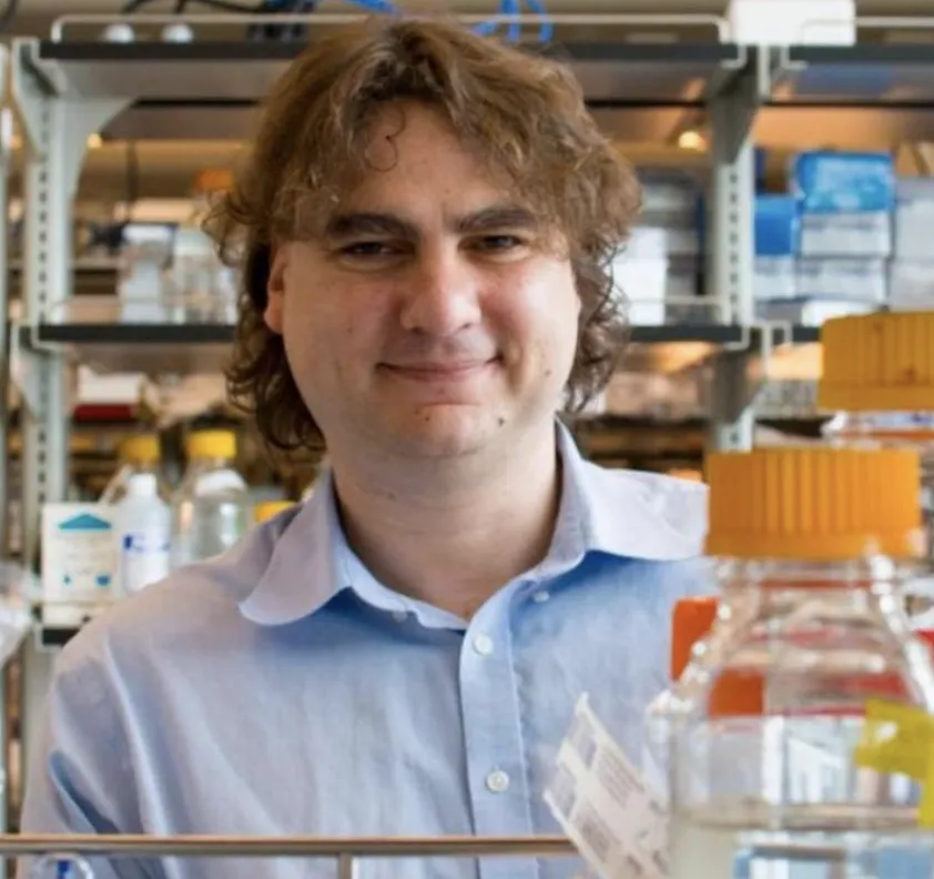 A portrait of Stavros standing behind a lab bench. He is wearing a light blue dress shirt, and there are glass pyrex bottles with orange caps in the foreground. 