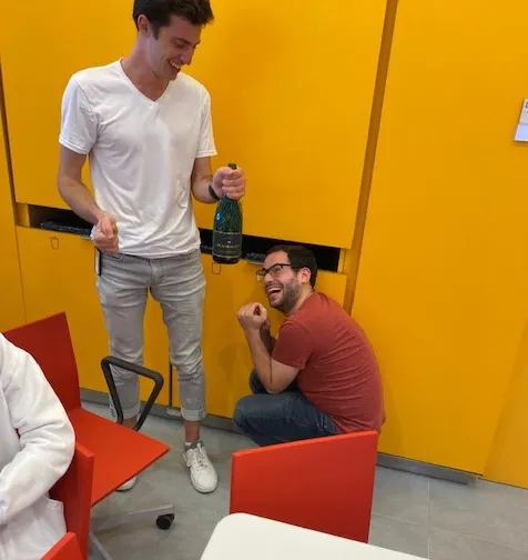Tibby wears a white tee, holds an opened champagne bottle in one hand and a cork in the other. Hani is smiling, looking up, still in a crouched position. 