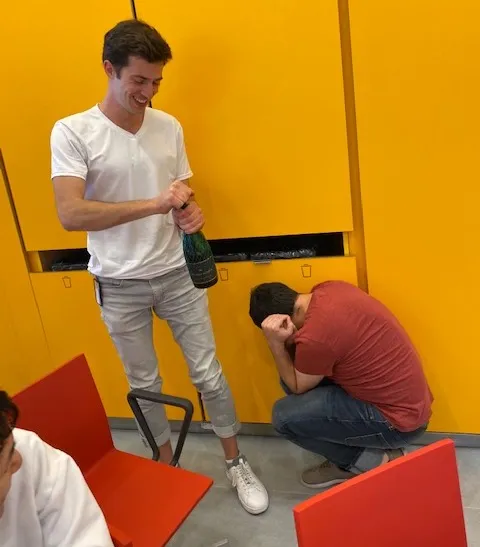 Tibby is wearing a white tee and rolled up jeans. He smiles while holding the champagne bottle. Hani is in a red tee and covers his ear as he huddles on the ground.
