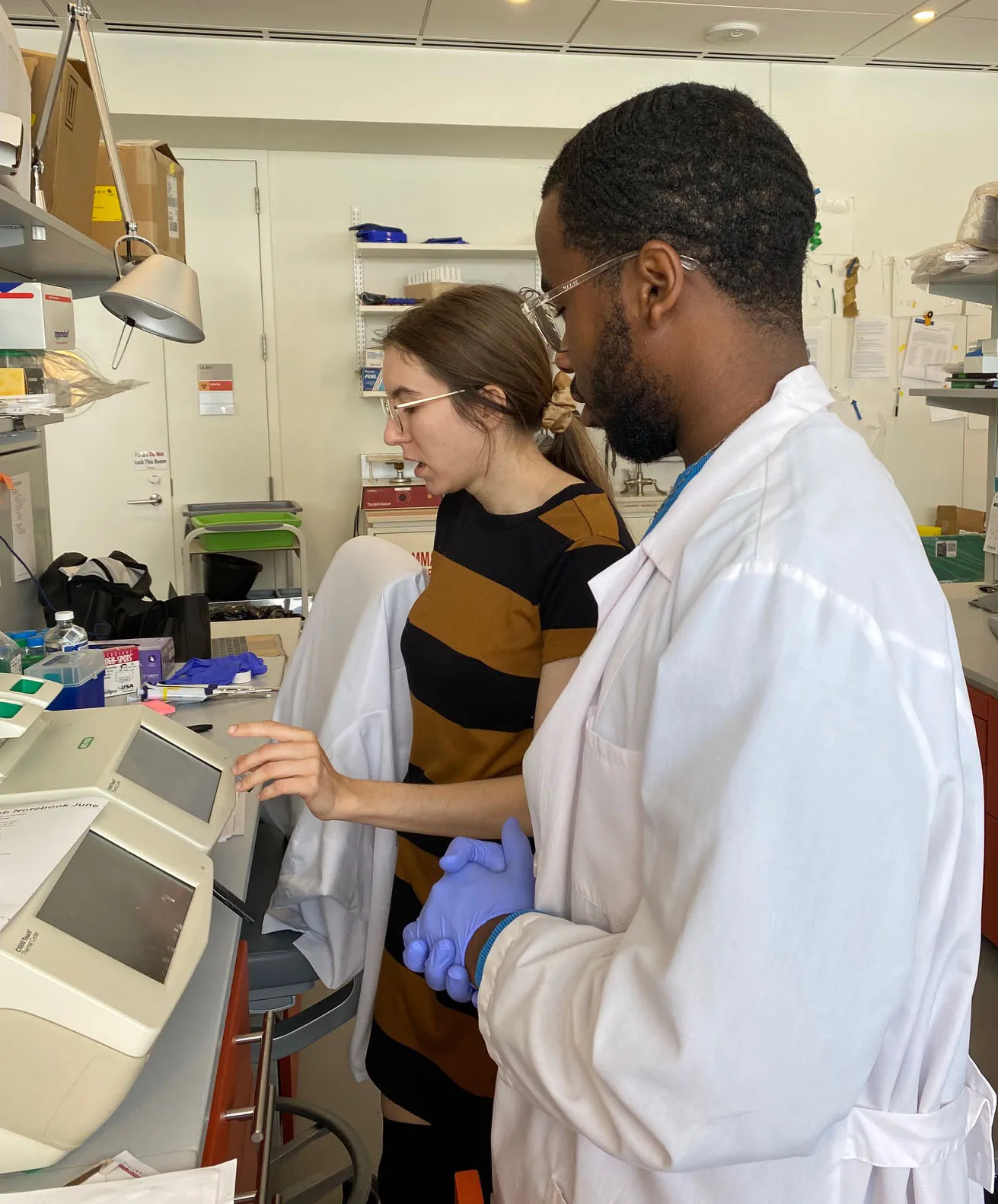 Chastin, in a lab coat and purple gloves, is being shown how to program the PCR machine by fellow lab member, Alexa.