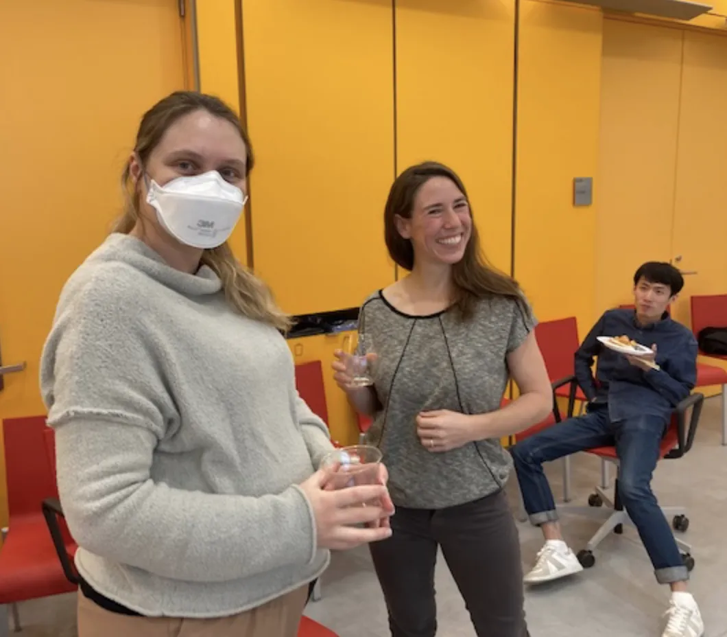Lisa is holding a soda, Rachel is standing and holding champagne, and Junming is seated and eating. 