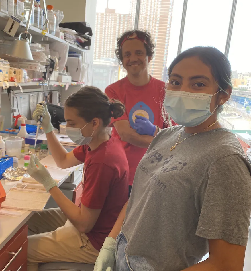 Aldo is pipetting into a tube at his bench, while America and Martin turn to smile at the camera. 