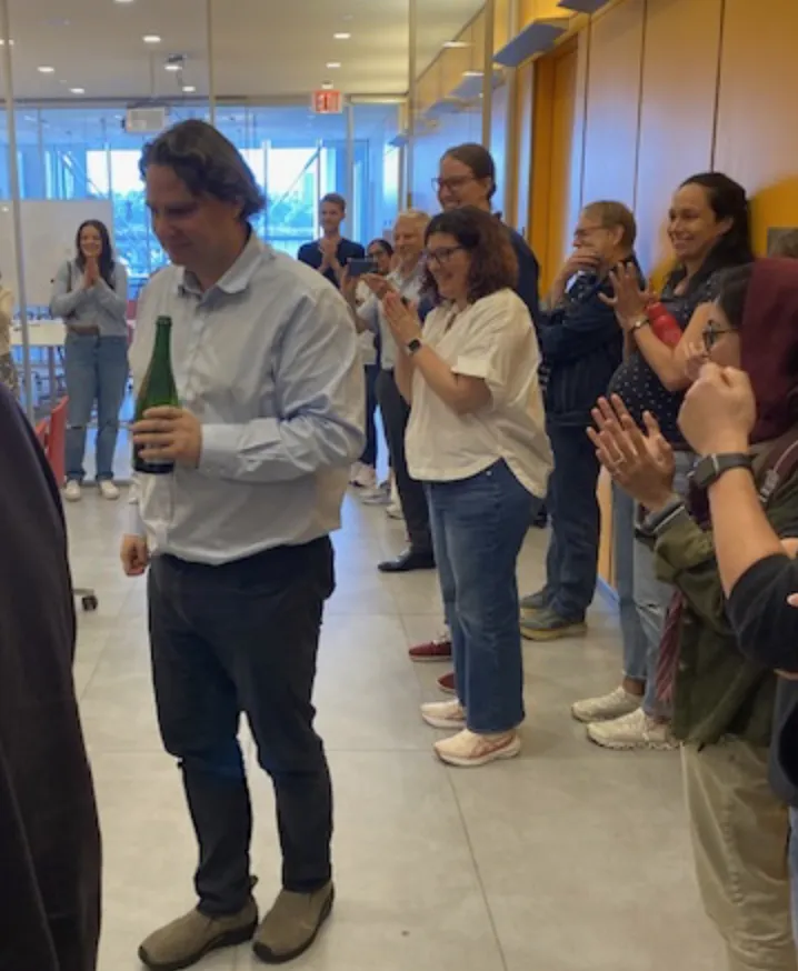 Someone holding a champagne bottle while a group gathers around clapping