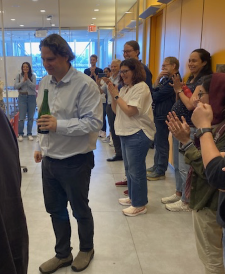Someone holding a champagne bottle while a group gathers around clapping