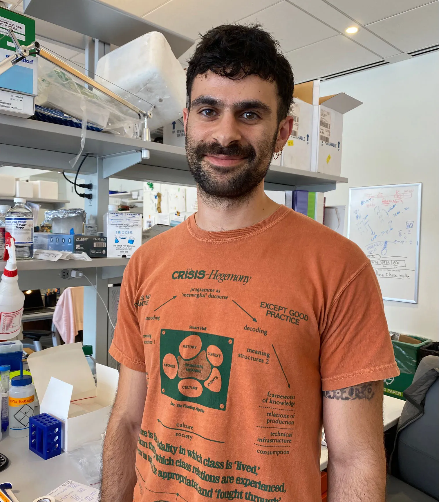 Ariel is smiling while wearing a rust colored tee and standing in front of his bench.