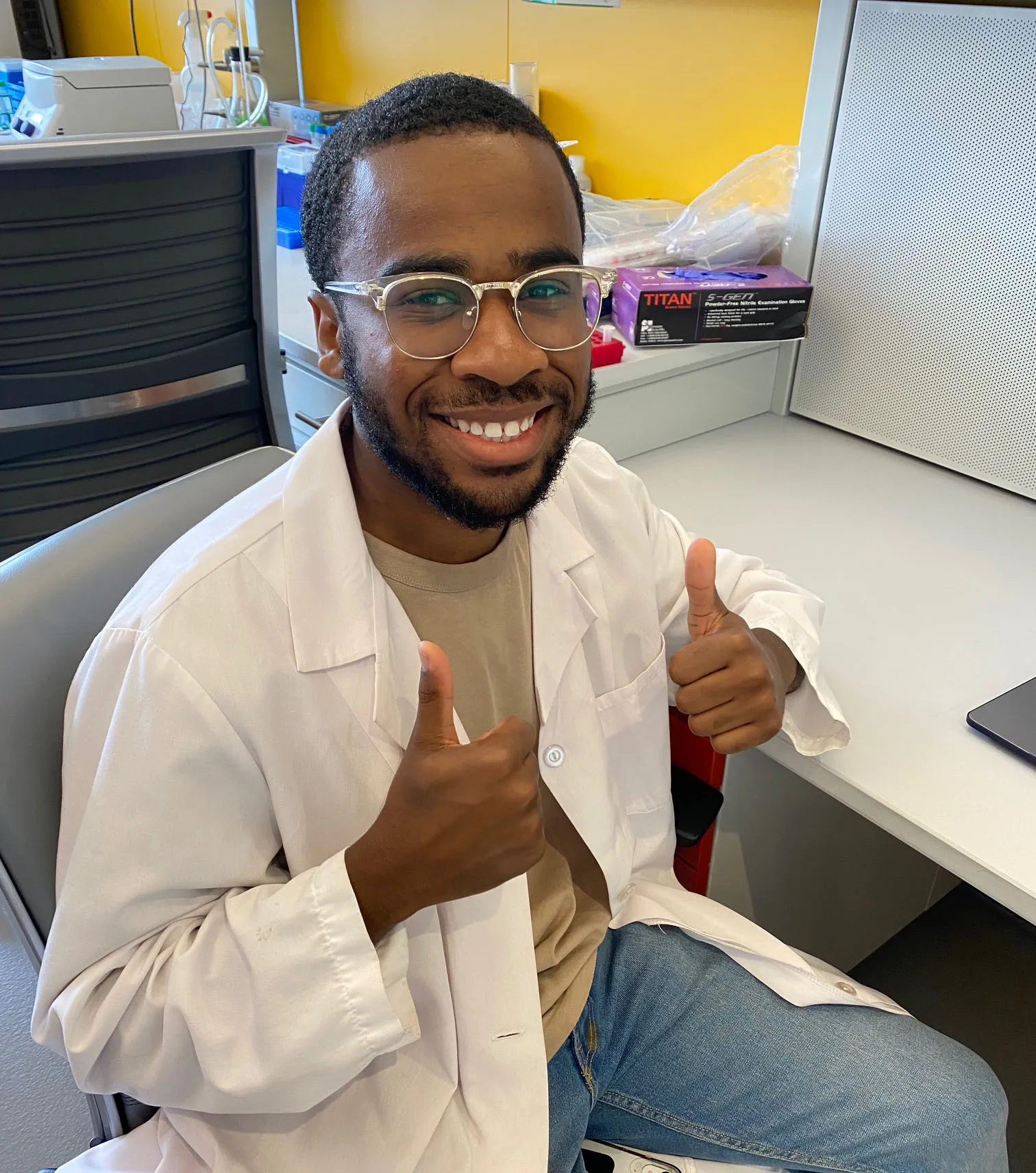 Chastin is smiling with his thumbs up, as he sits at the desk next to his bench. 