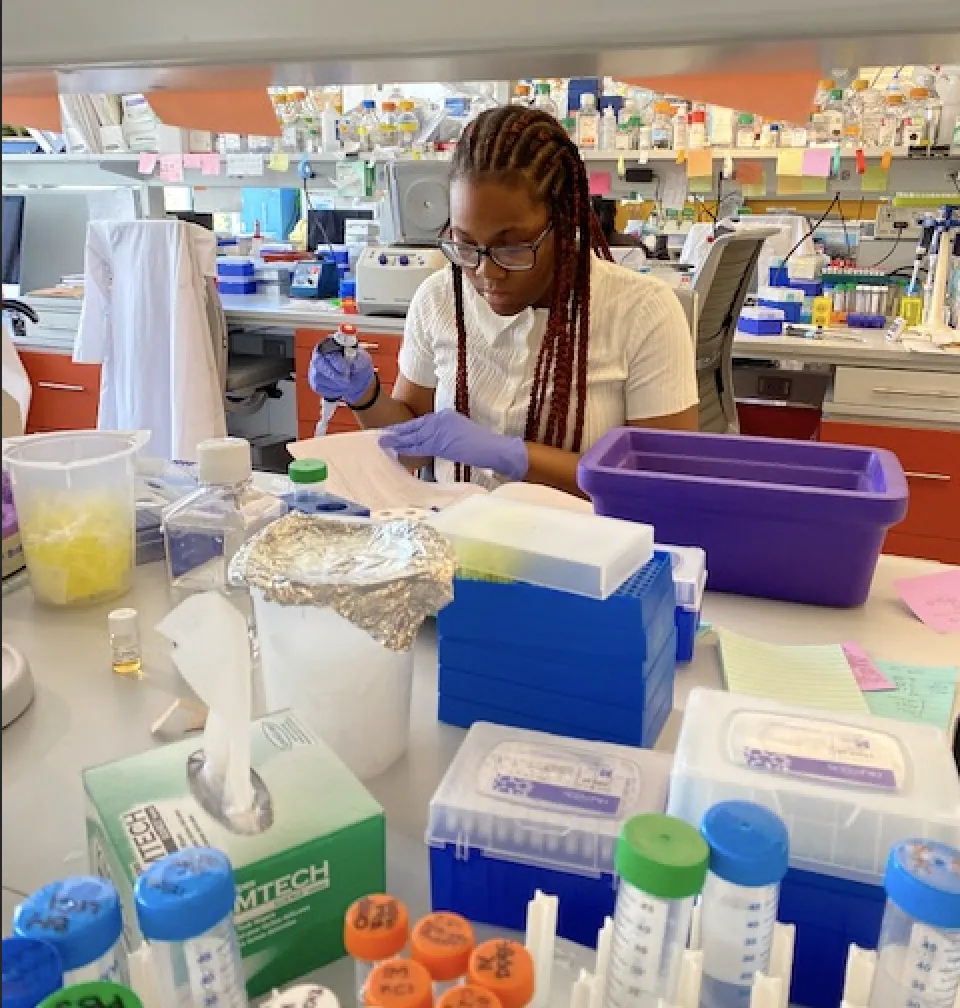Joanne is wearing purple nitrile gloves while holding a pipettor. She works independently at the bench while reading a protocol book, surrounded by tubes and a purple  ice bucket. 